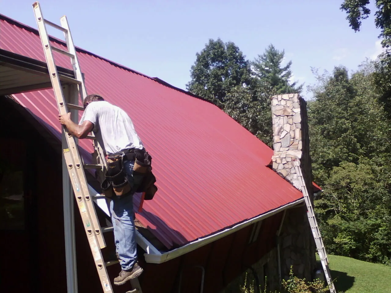 Roofer on a ladder beside a red metal roof and stone chimney — Soffit & Fascia Repair by Roofing Brothers in Sidney